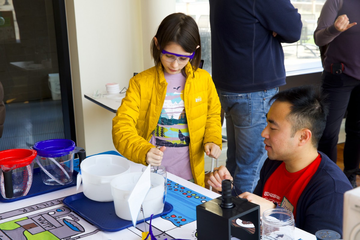 A young girl wearing a bright yellow jacket and protective glasses engages with a staff member at a booth.