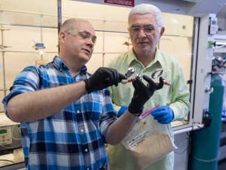 Man wearing black rubber gloves holding a piece of lab equipment as man holding plastic bag of wood chips looks on
